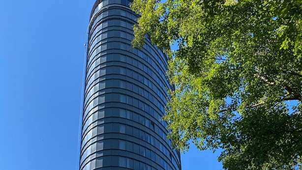 A tall, cylindrical glass building with a curved facade, surrounded by green foliage.