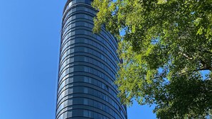 A tall, cylindrical glass building with a curved facade, surrounded by green foliage.