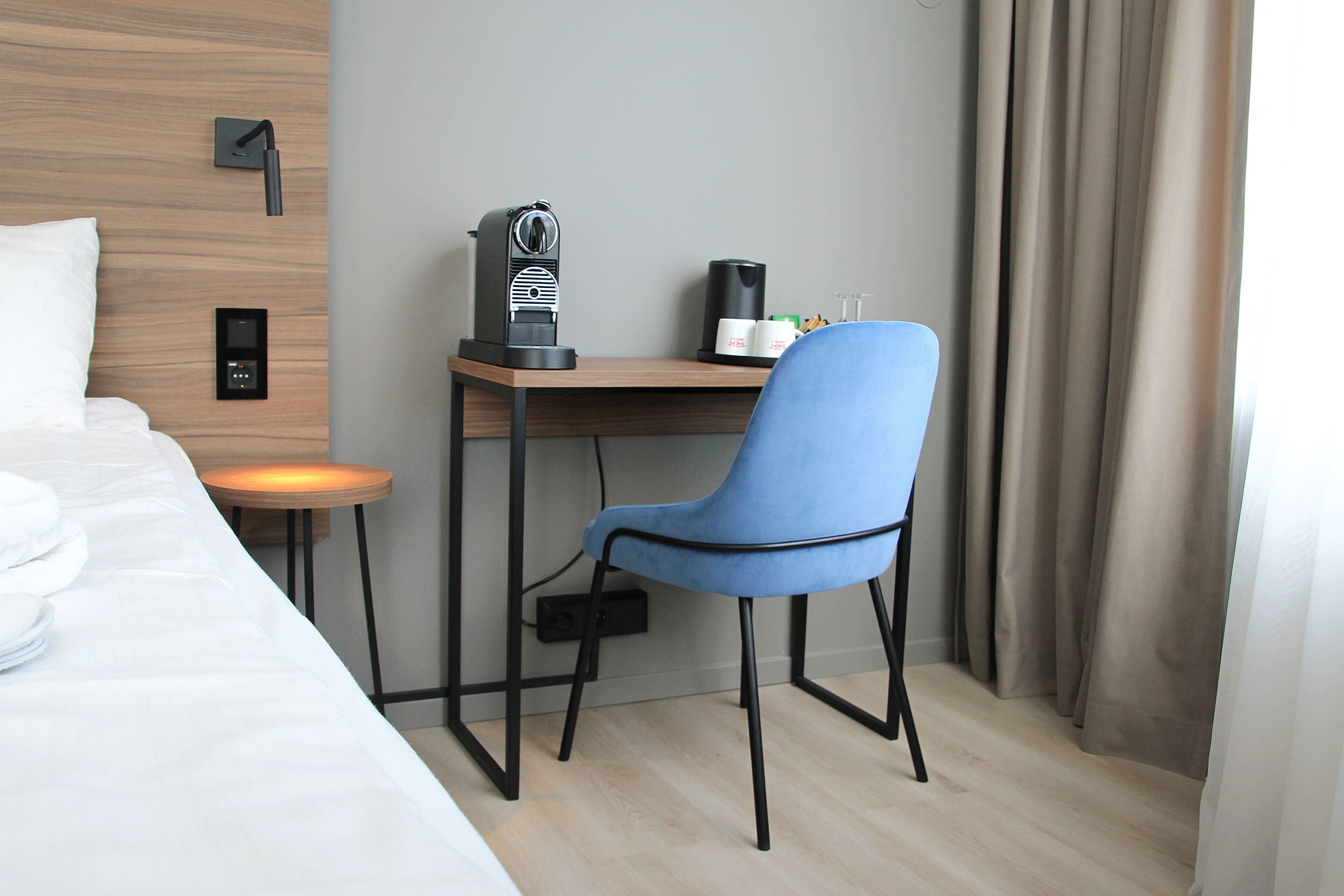 A hotel room with a bed, a desk with a coffee maker, a chair, and a small table with a lamp.