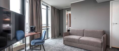 A hotel room with a large window, a sofa, a desk with a chair, and a television.