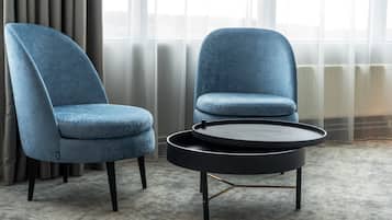 Duas poltronas azuis e uma mesa de centro redonda preta num quarto com piso coberto de carpete e janela com cortinas leves.
