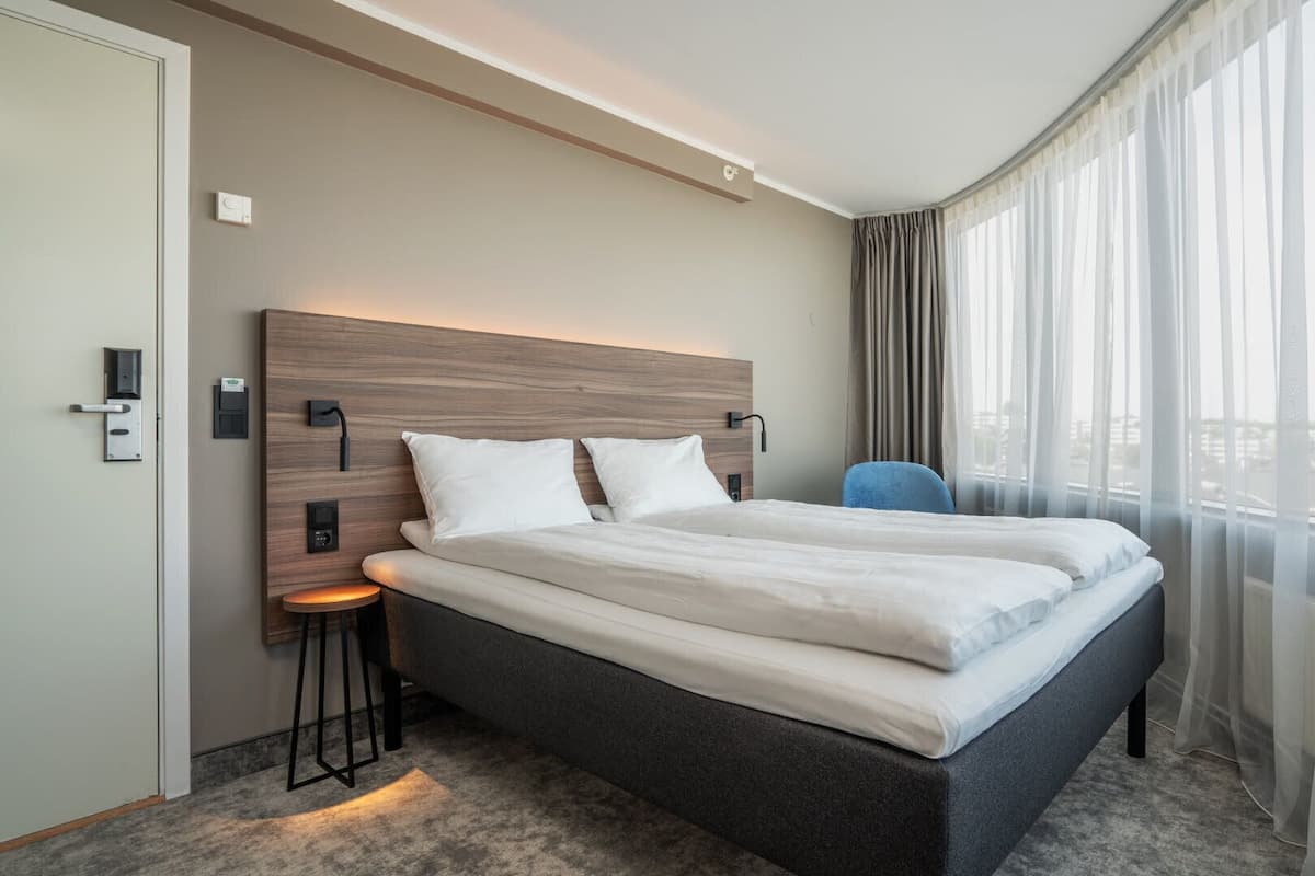 A modern hotel room with a large bed, a nightstand, a chair, and a view of the city through sheer curtains.