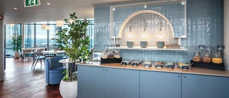 A modern kitchen area with a buffet setup, including various food containers and a large potted plant.