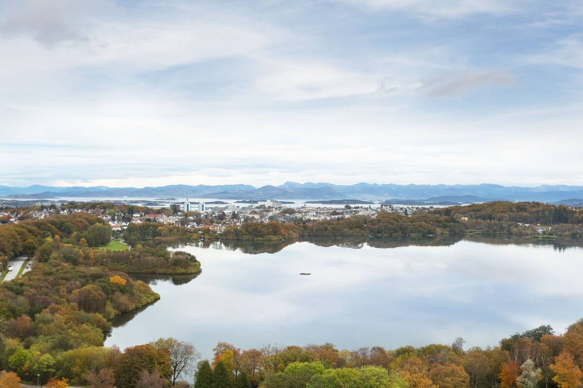A panoramic view of a lake surrounded by autumn-colored trees and a distant cityscape.