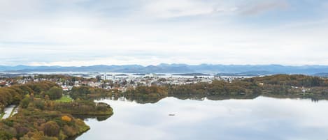 Vista panorâmica de um lago rodeado por árvores de cores outonais e uma paisagem urbana ao longe.