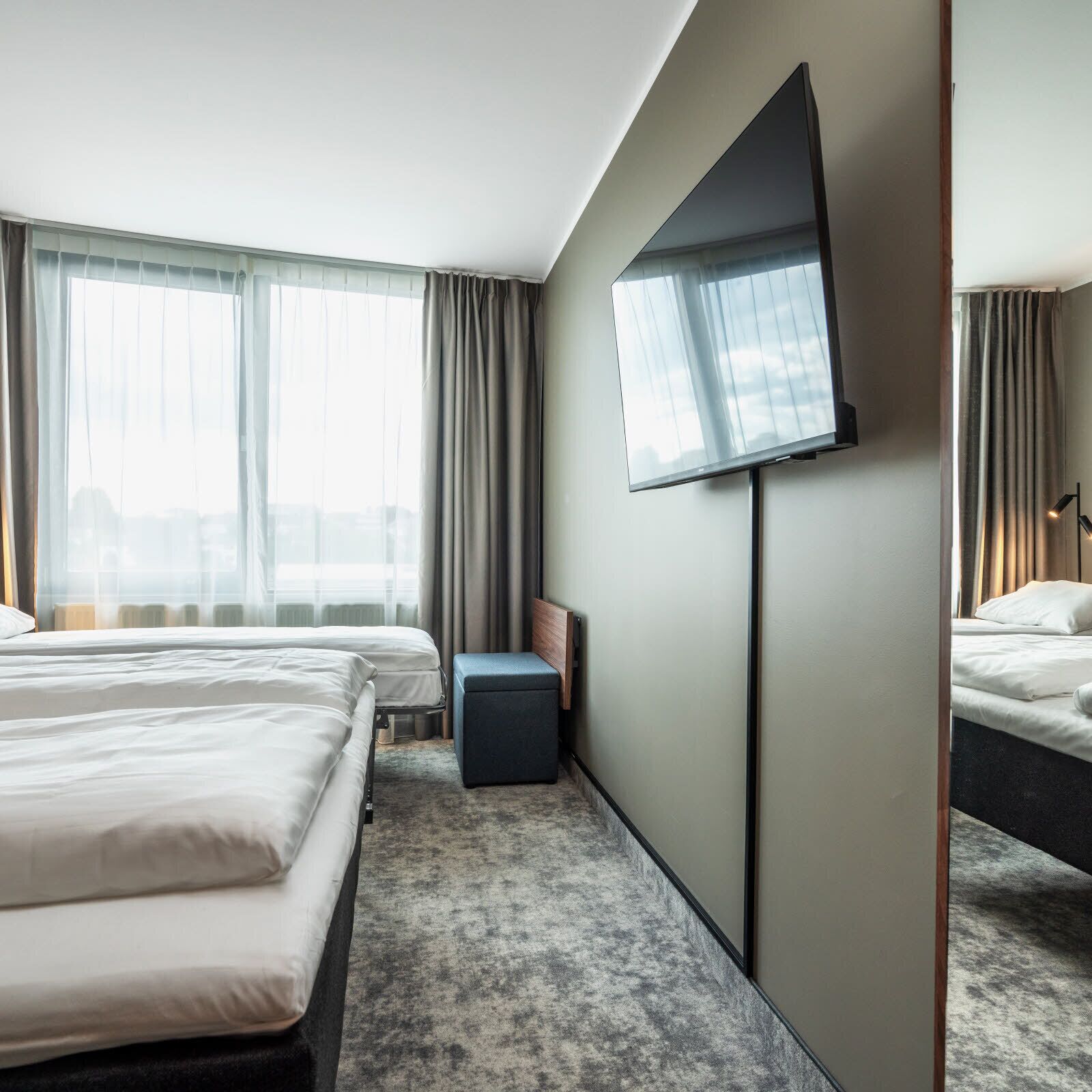 Hotellihuone, jossa on kaksi sänkyä, seinälle asennettu taulutelevisio, pieni oleskelualue ja suuret ikkunat, joissa on verhot.