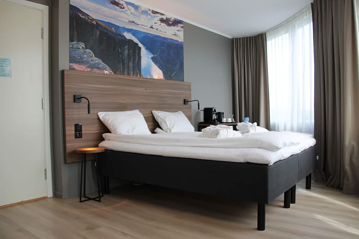 A modern hotel room with a large bed, a nightstand, a coffee maker, and a painting of a mountain landscape.