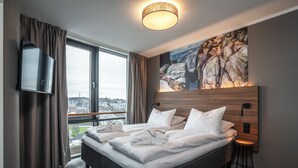 A hotel room with a large bed, a TV, a painting of rocks, and a view of a cityscape.