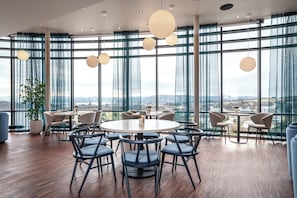 A modern dining area with round tables and chairs, large windows with a view of the city, and hanging spherical light fixtures.