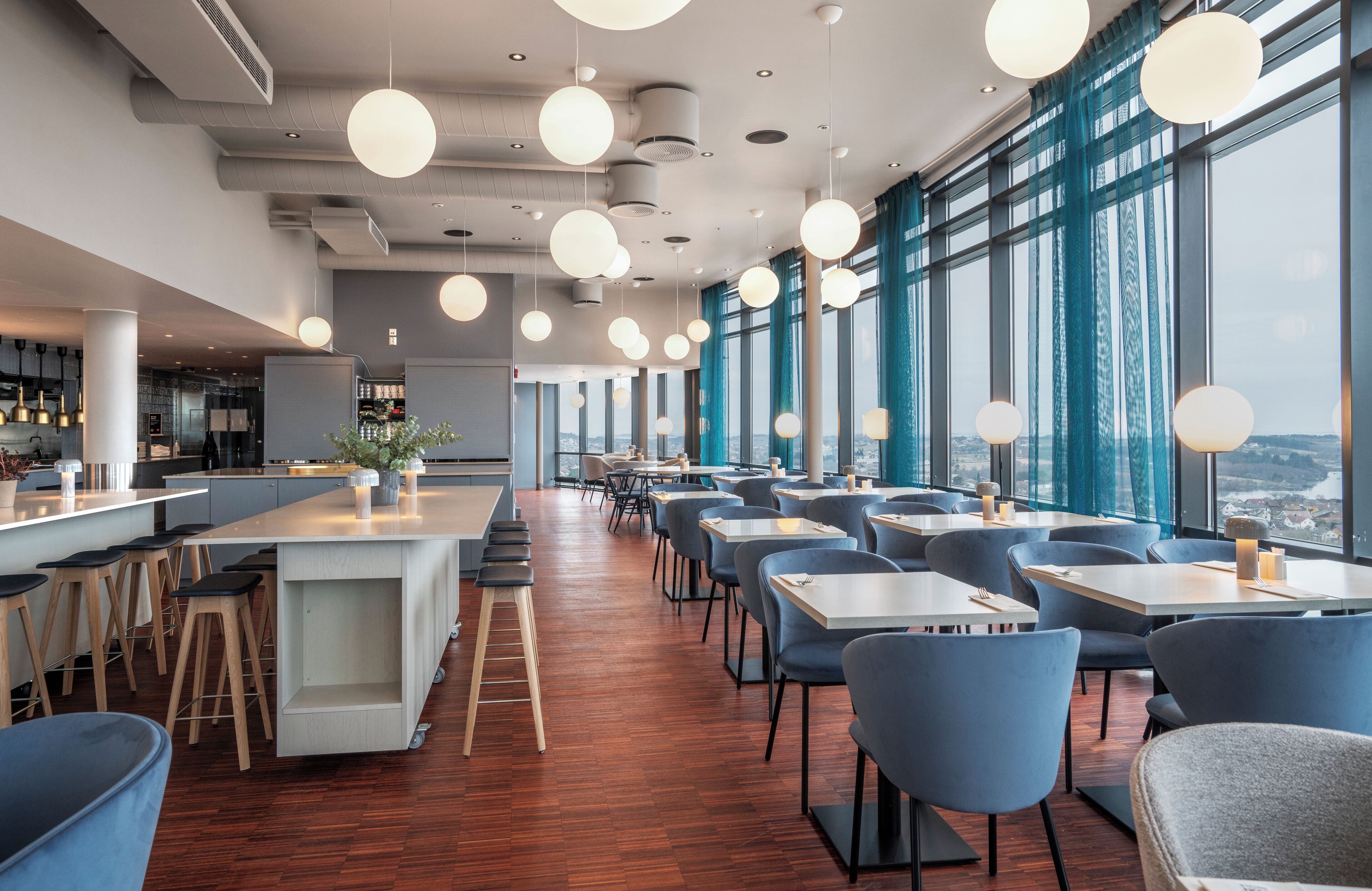 A modern restaurant with high ceilings, pendant lighting, and large windows offering city views.