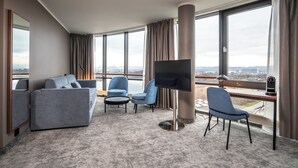 A modern hotel room with a large window, a flat-screen TV, a sofa, two armchairs, and a desk with a lamp.