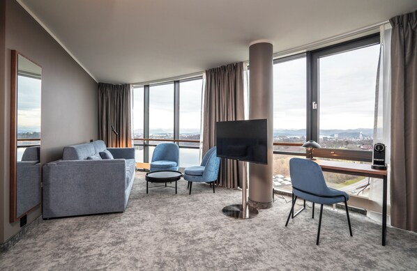 Una habitación de hotel moderna con un ventanal grande, un televisor de pantalla plana, un sofá, dos sillones y un escritorio con lámpara.