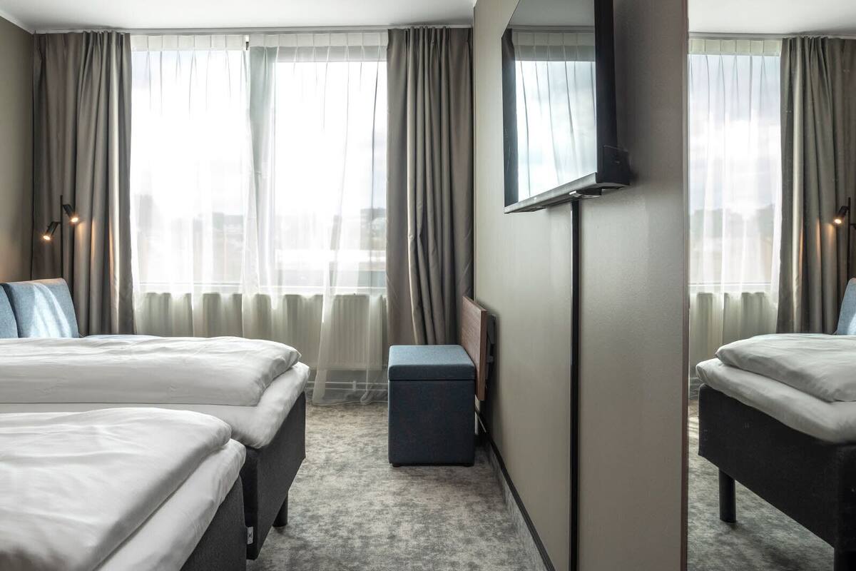 A hotel room with two beds, a large mirror, and a view of the city through sheer curtains.