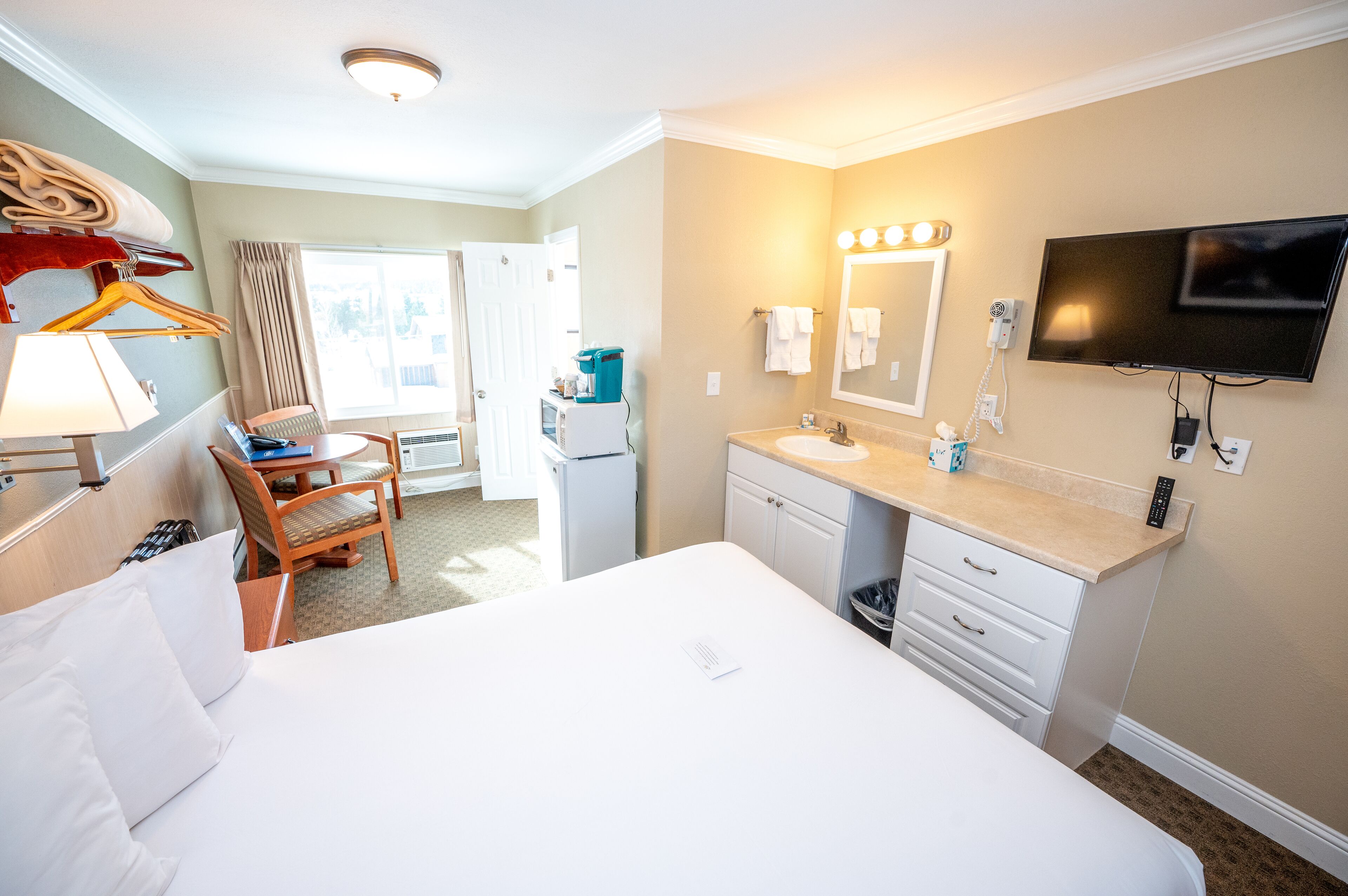 A hotel room with a bed, a desk, a chair, a desk lamp, a TV, a mirror, a sink, and a window with curtains.