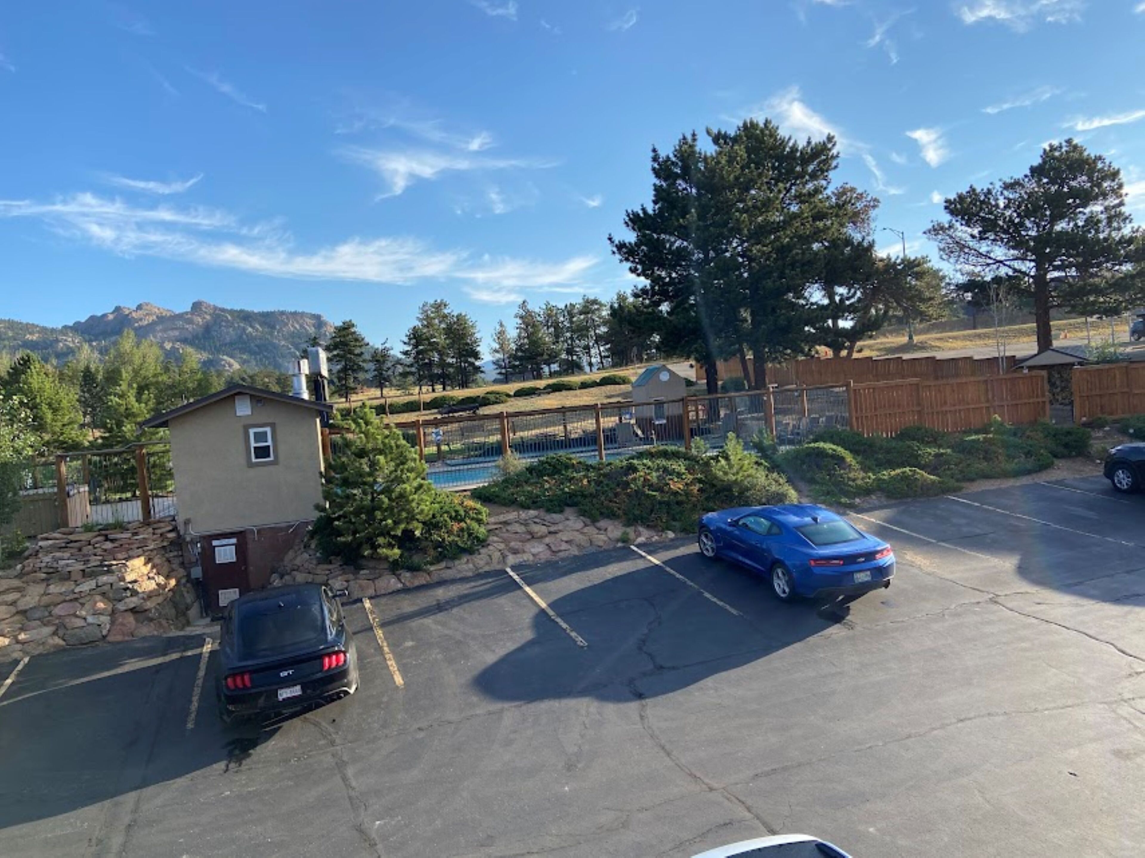A parking lot with two cars, a small building, and a fenced area with a pool.