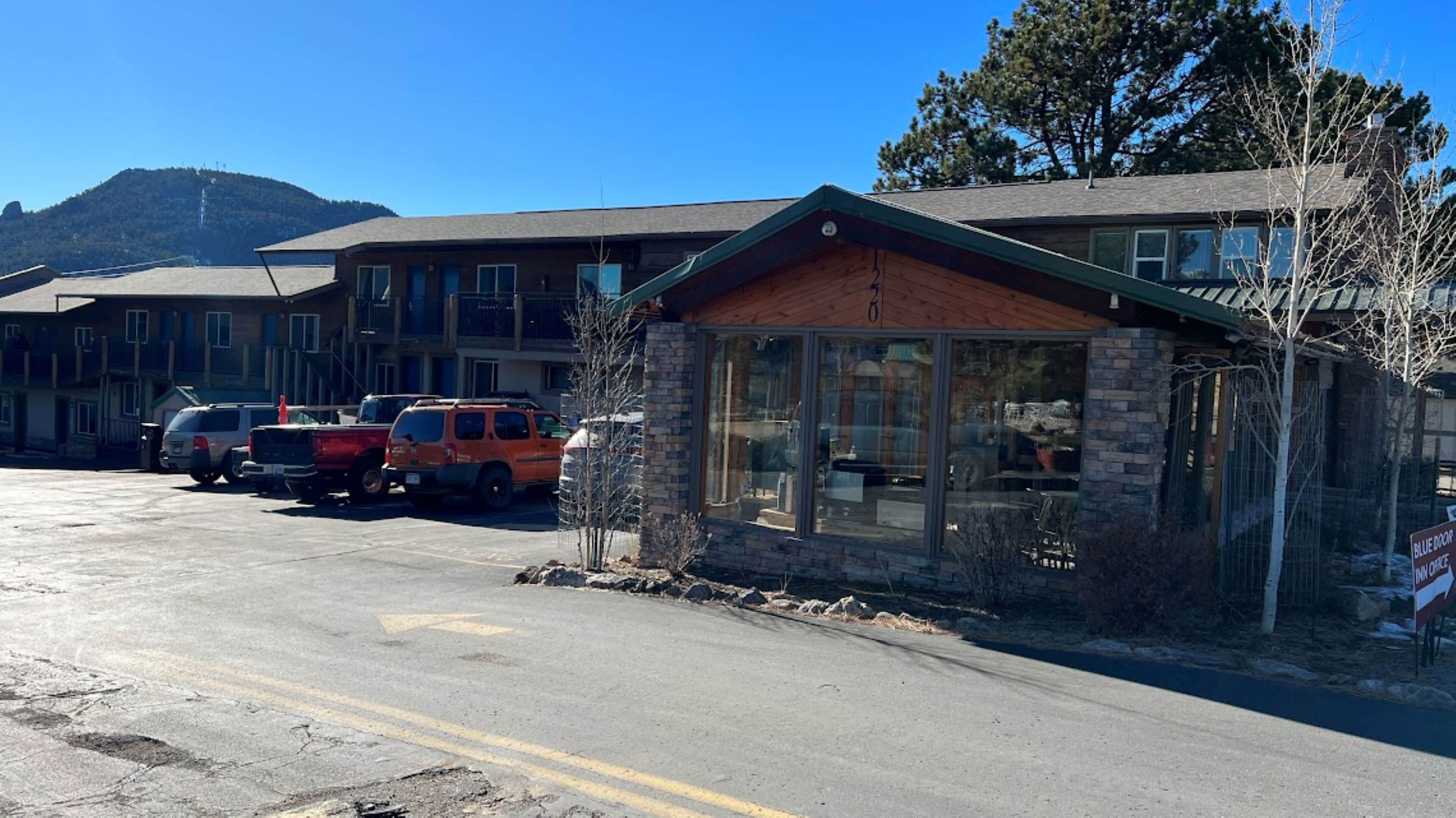 Un motel con fachada de piedra, coches aparcados y una montaña al fondo.