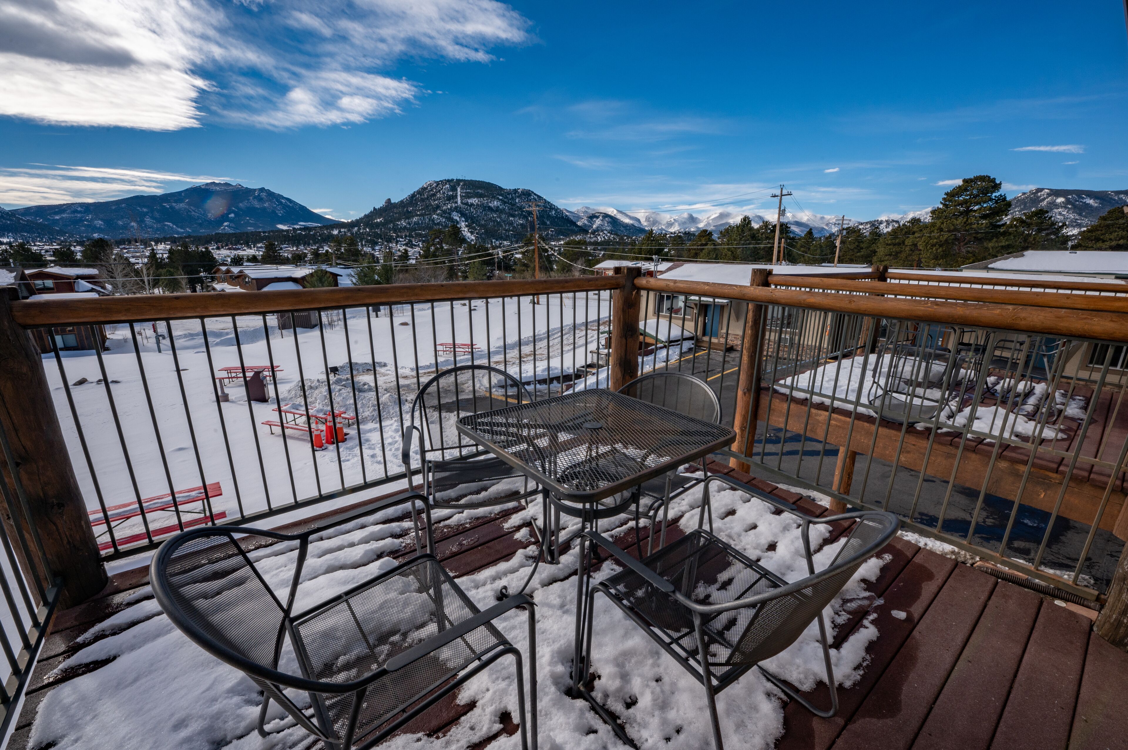 A balcony with metal chairs and tables, a wooden railing, and a snowy landscape with mountains in the background.