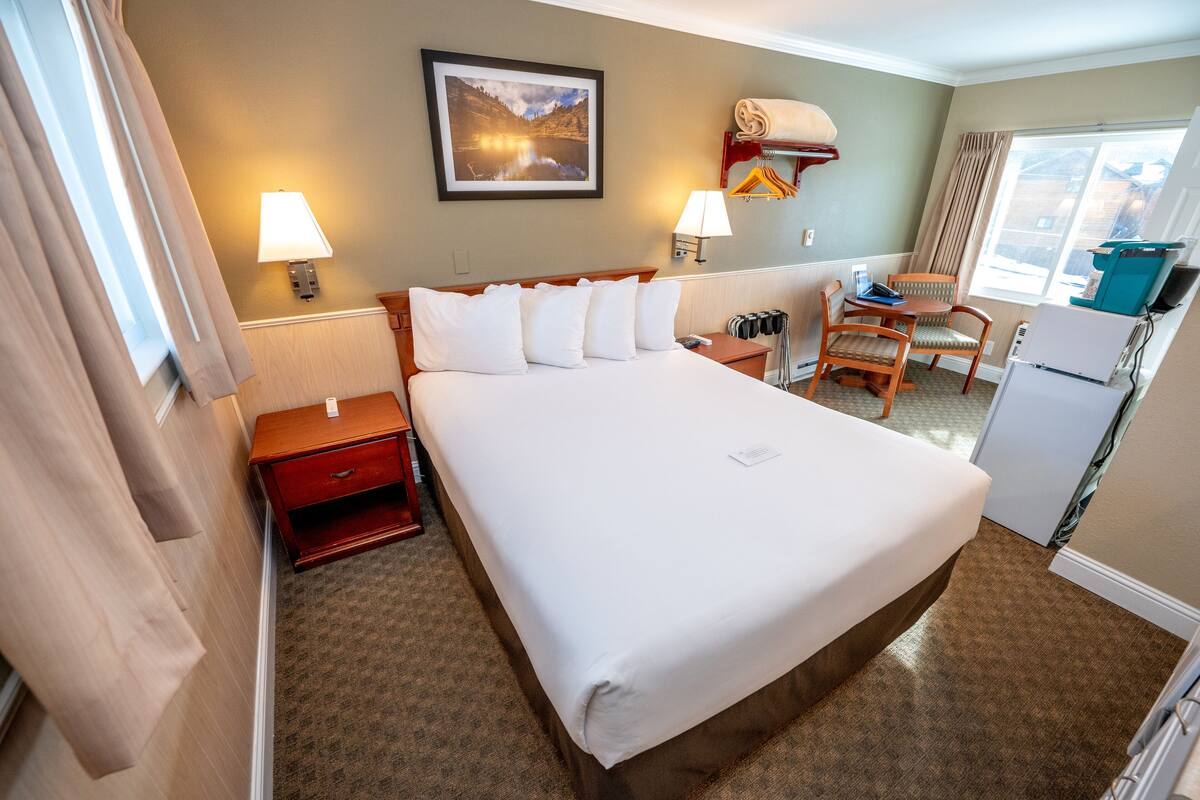 Une chambre d’hôtel avec un grand lit, des tables de chevet, un bureau avec une chaise, une lampe, un tableau au mur et une fenêtre avec des rideaux.