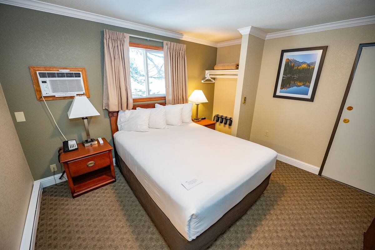 Une chambre d’hôtel avec un grand lit, une table de chevet avec une lampe, un climatiseur fixé au mur, une fenêtre avec des rideaux et un tableau accroché au mur.