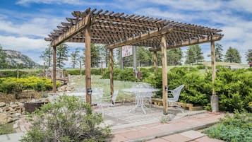 Una pérgola de madera con mesa y sillas debajo, rodeada de vegetación y un paisaje montañoso.