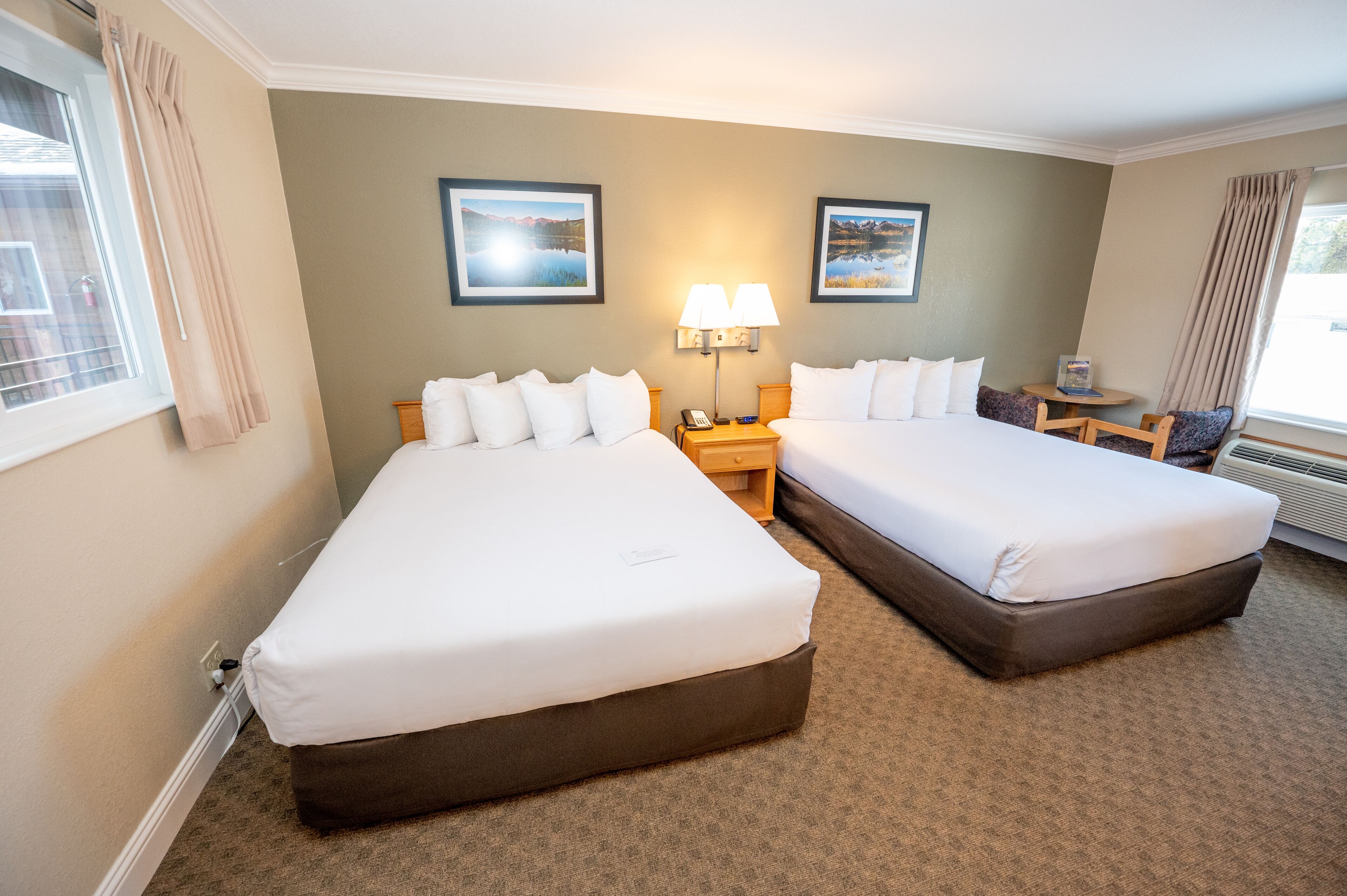 Habitación de hotel con dos camas, una mesita, una lámpara y dos cuadros en la pared.