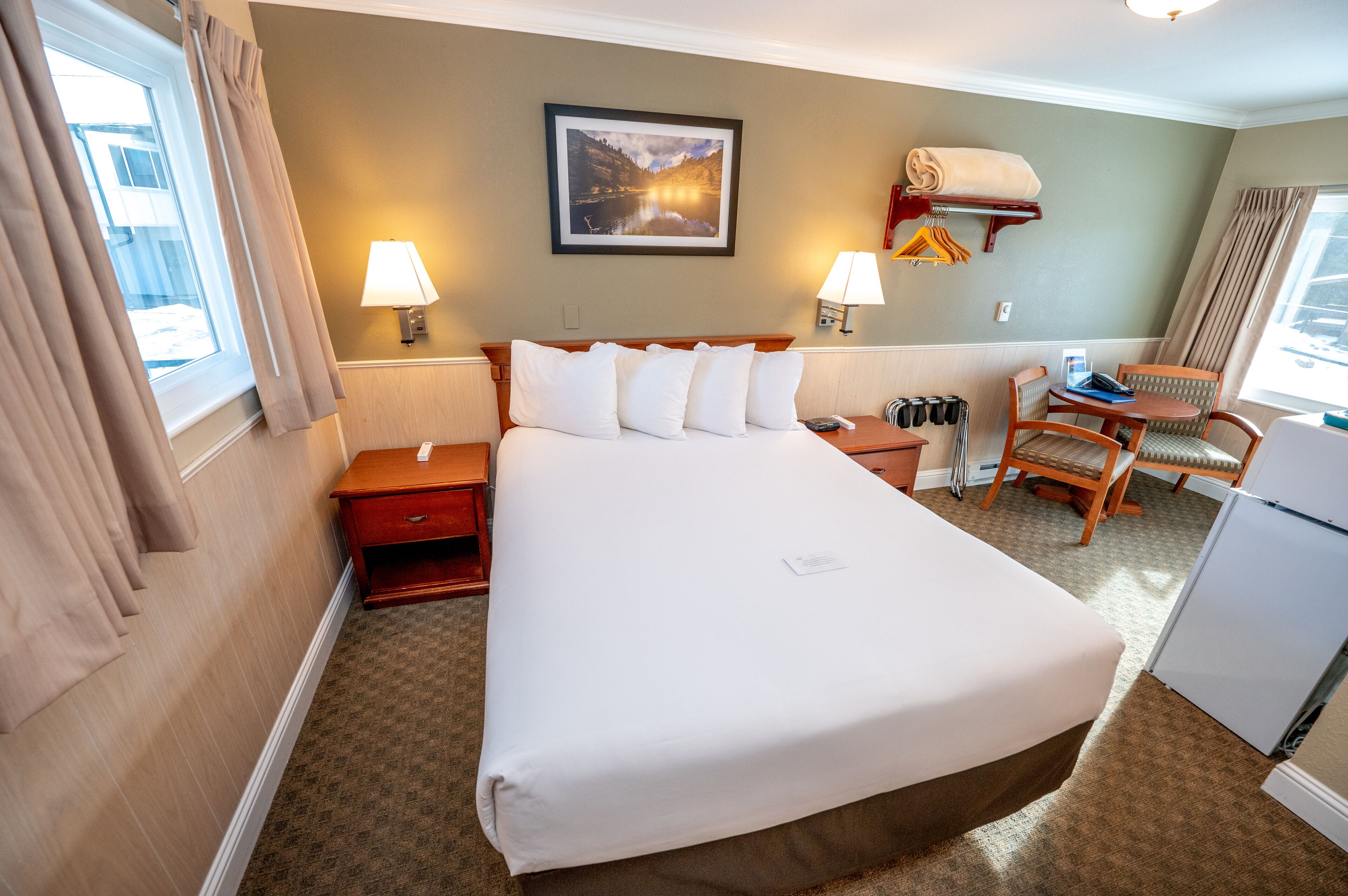 Habitación de hotel con una cama grande, dos mesitas de noche, un escritorio con silla, una lámpara de pared, un cuadro y un refrigerador pequeño.