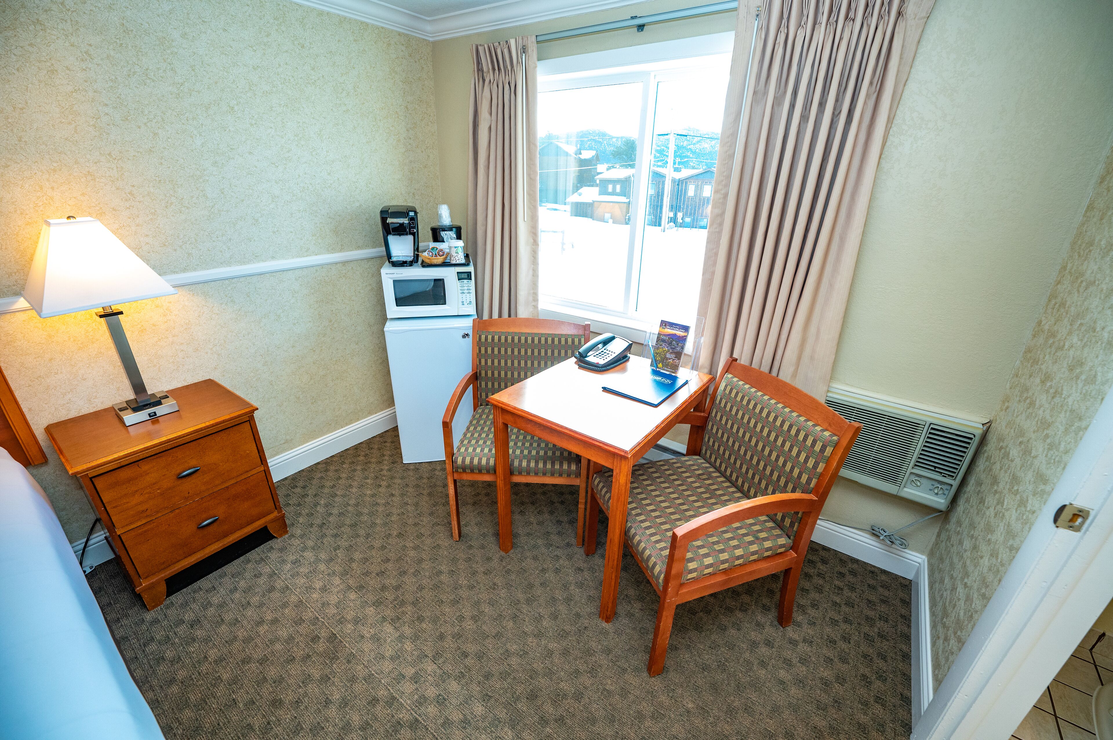 A small hotel room with a single bed, a wooden nightstand with a lamp, a table with chairs, a microwave, a telephone, and a window with curtains.
