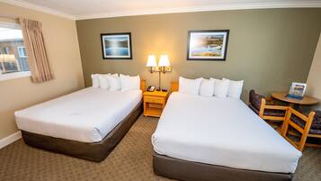A hotel room with two single beds, a small table, a chair, a lamp, and two framed pictures on the wall.