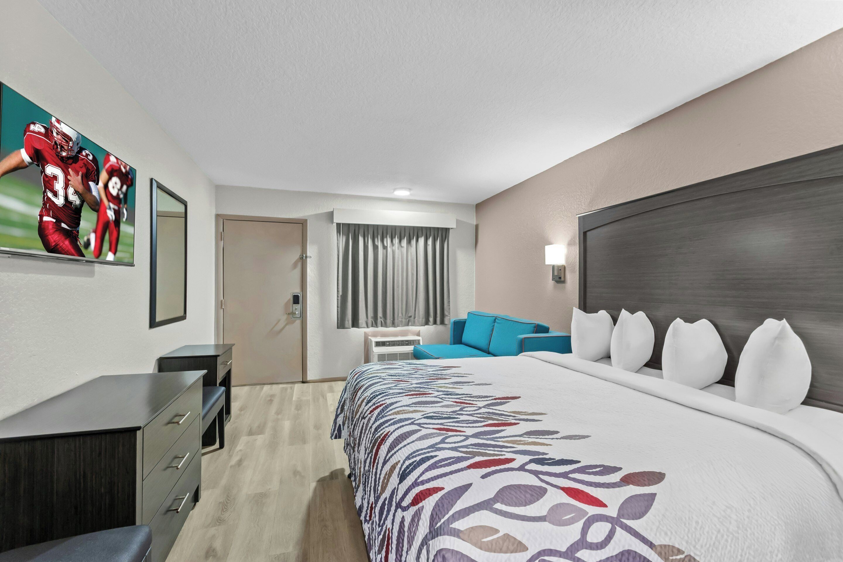Habitación de hotel con una cama grande, un escritorio con cajones, una silla, un sofá, una ventana con cortinas y un cartel deportivo en la pared.