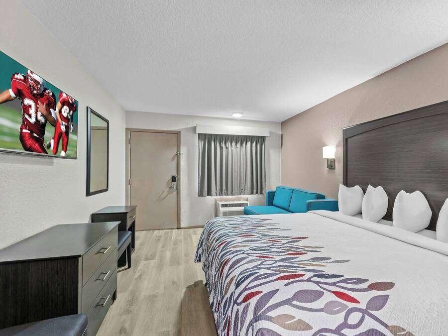 A hotel room with a large bed, a desk with drawers, a chair, a sofa, a window with curtains, and a wall with a sports poster.