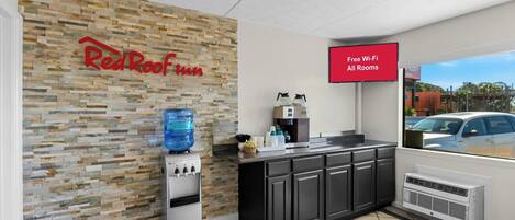 A RedRoof Inn reception area with a stone wall, a water dispenser, a coffee machine, and a sign indicating free Wi-Fi in all rooms.