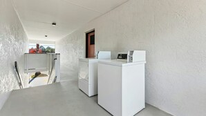 Un área de lavandería con dos lavadoras blancas y una secadora, una ventana pequeña con vistas, y una puerta que da a otra habitación.