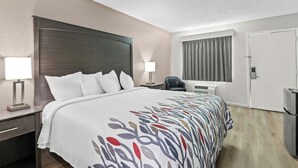 A hotel room with a large bed, bedside lamps, a desk, a chair, an air conditioning unit, and a door.