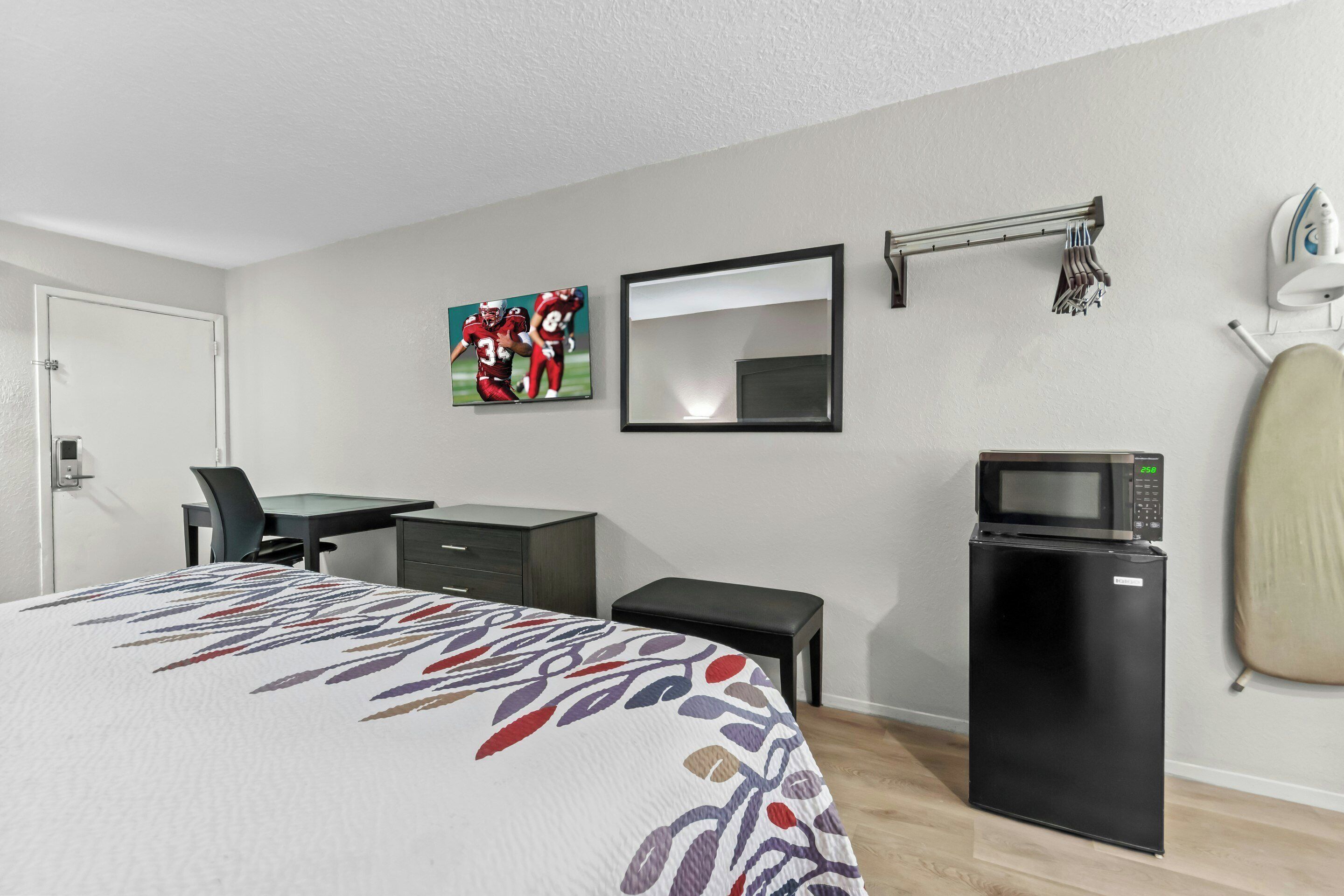 A hotel room with a bed, a desk, a chair, a small black refrigerator, a microwave, a mirror, and a towel rack.