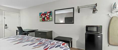 A hotel room with a bed, a desk, a chair, a small black refrigerator, a microwave, a mirror, and a towel rack.