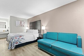 A modern hotel room with a bed, a sofa, a nightstand, and a wall-mounted TV. - Red Roof Inn Panama City Beach (Panama City Beach)