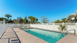 A rectangular swimming pool with clear blue water, surrounded by a white fence and palm trees.