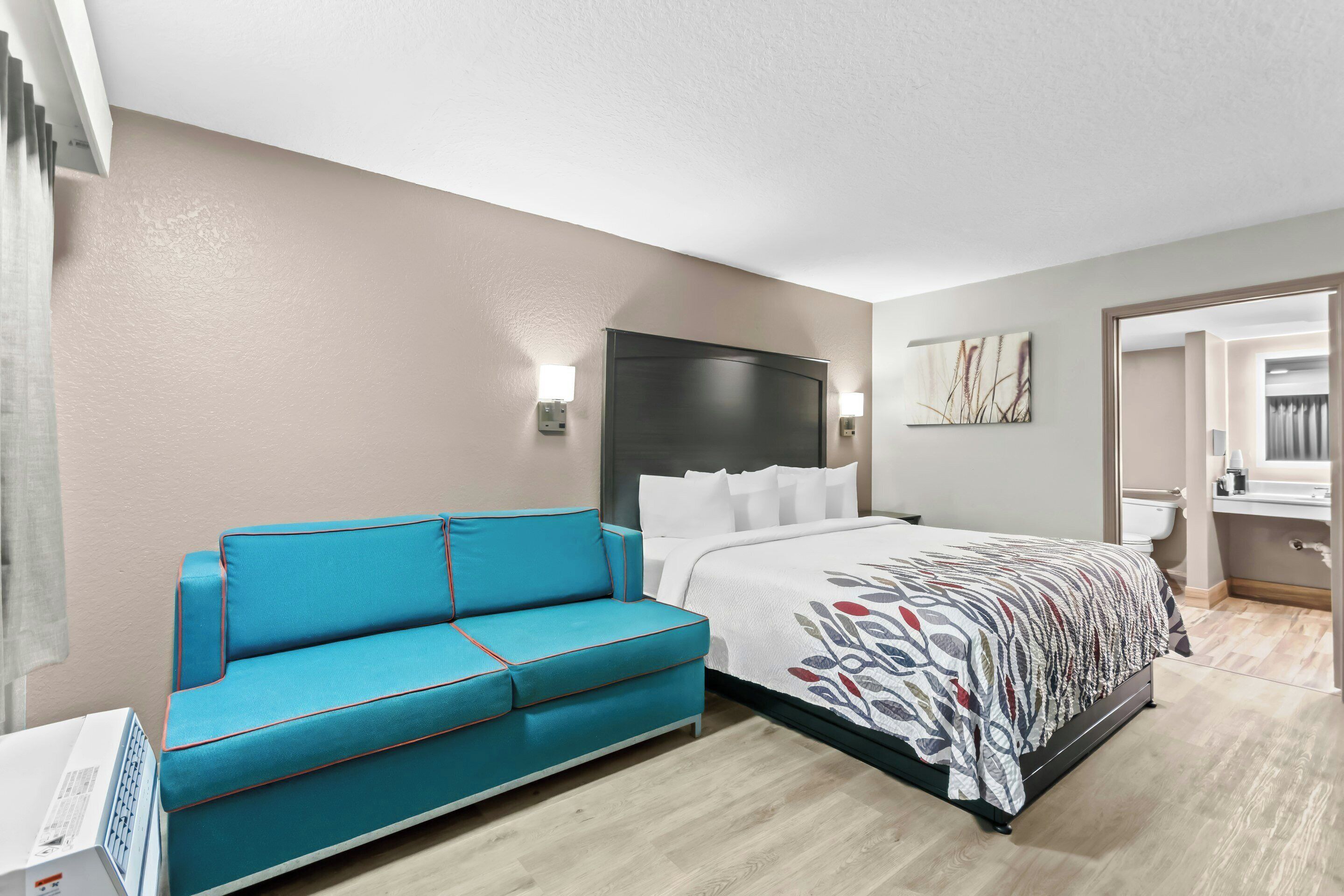 Quarto de hotel moderno com uma cama, um sofá azul, uma mesa de cabeceira e um banheiro visível por uma porta aberta.