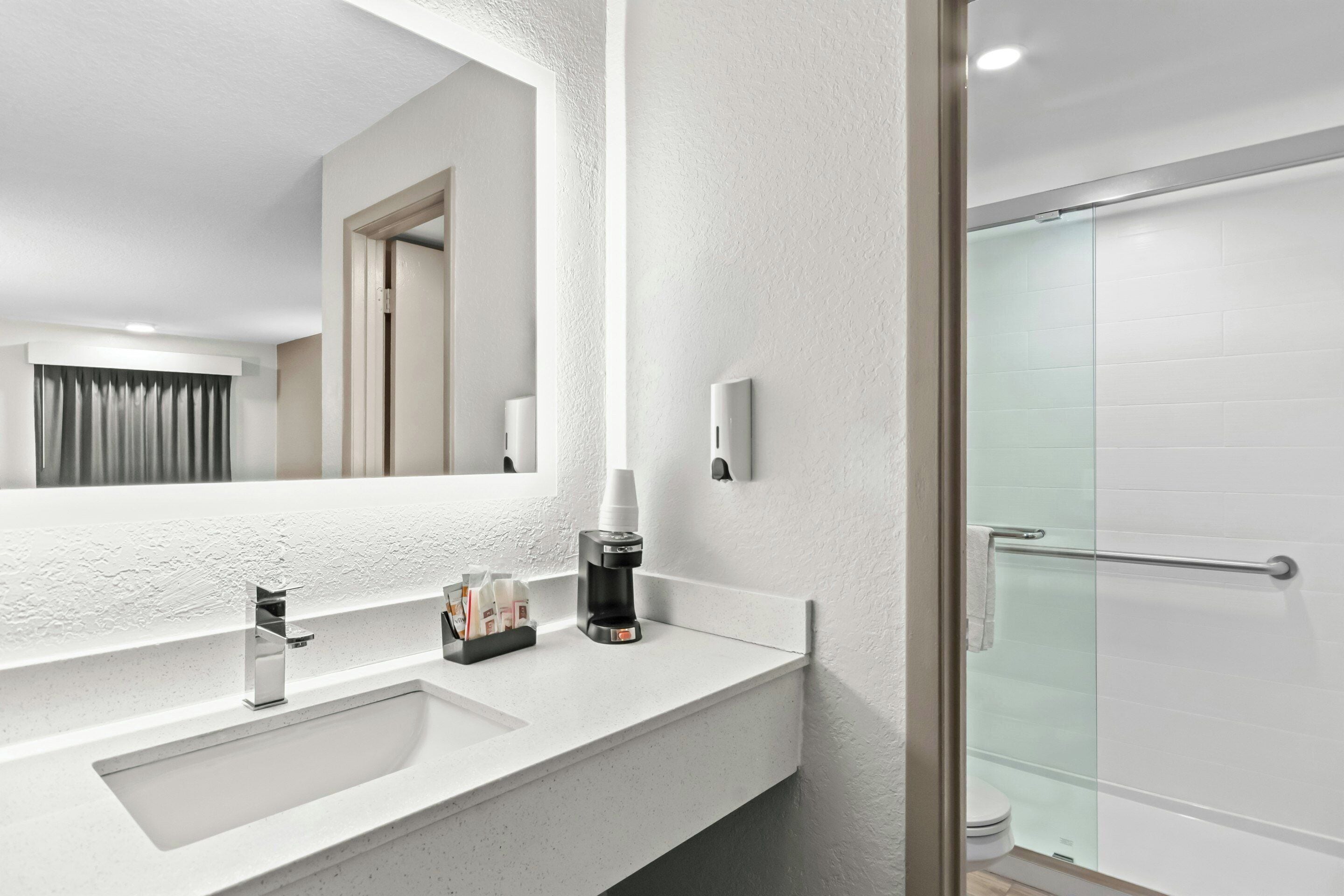 A modern bathroom with a large mirror, a sink, a coffee maker, and a glass shower enclosure.