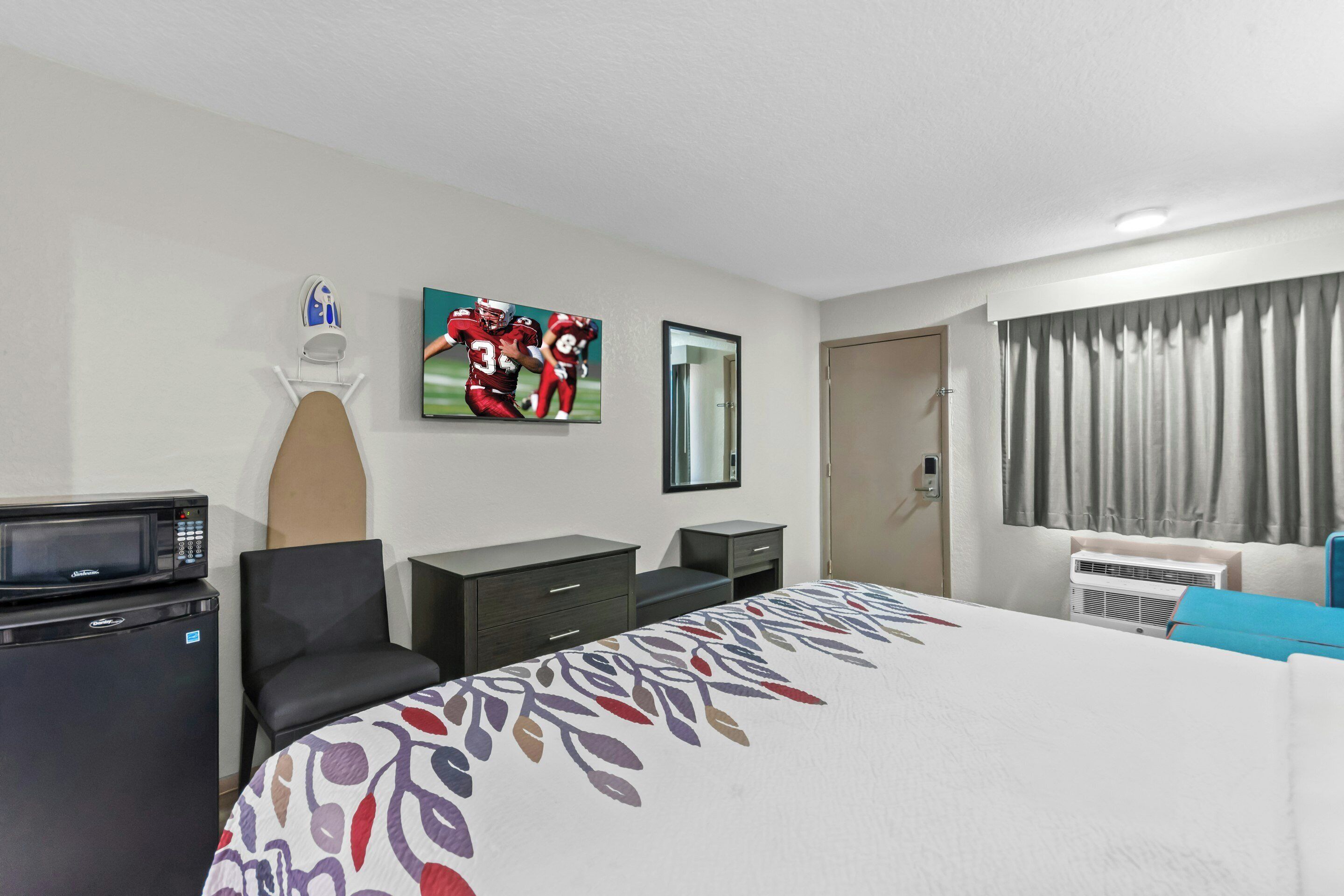 A hotel room with a bed, a desk, a chair, a microwave, a small refrigerator, a TV, and a window with curtains.