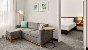 A modern hotel room with a grey sectional sofa, a round side table, a bed with white bedding, and two abstract wall art pieces. - TownePlace Suites by Marriott Chicago Lombard (Lombard)