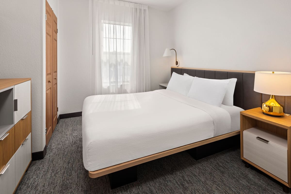 A hotel room with a bed, a nightstand with a lamp, a wardrobe, and a window with sheer curtains.