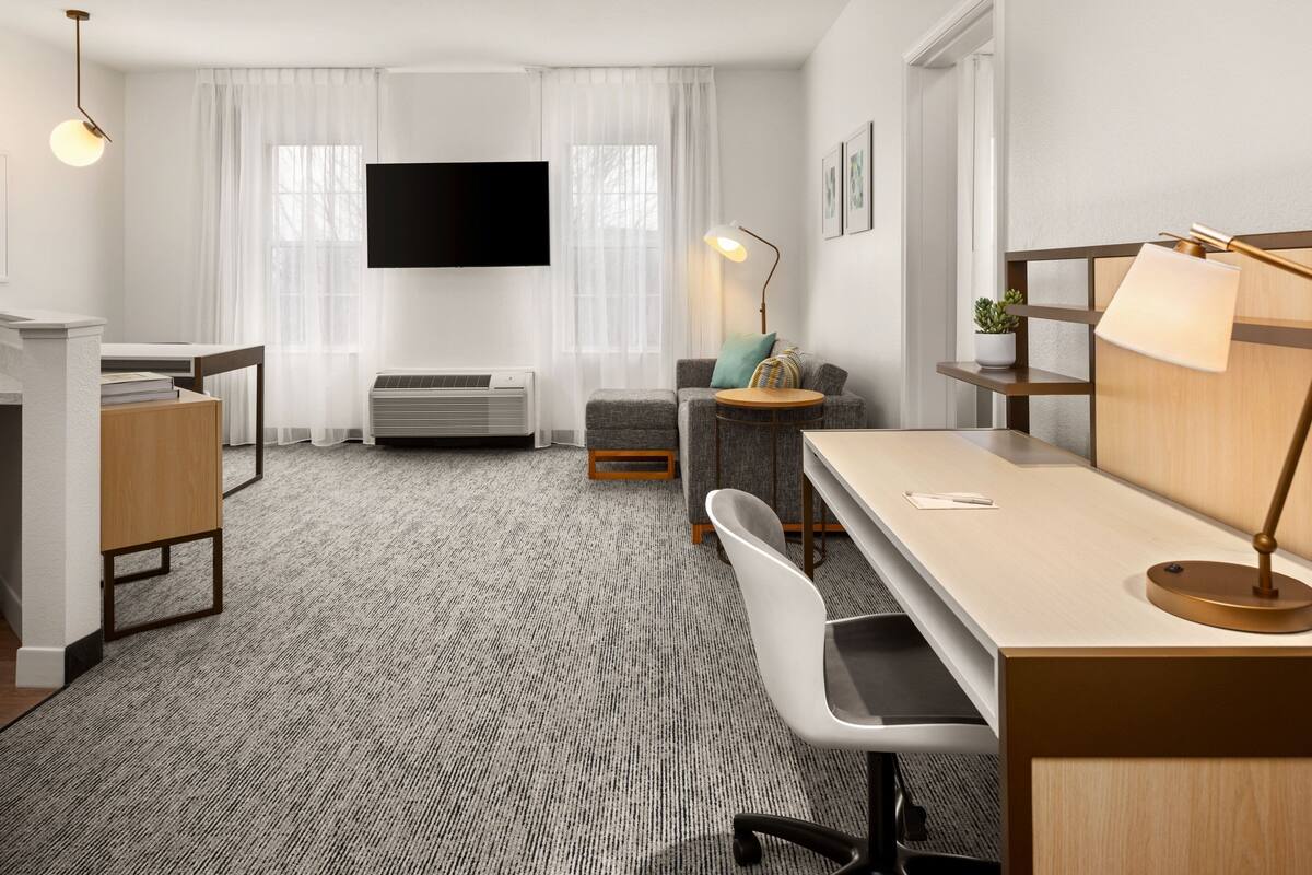 A modern hotel room with a flat-screen TV mounted on the wall, a desk with a lamp, a comfortable sofa, and a small side table.