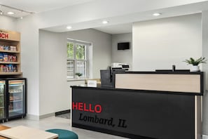A hotel reception area with a black reception desk labeled "HELLO Lombard, IL," a refrigerator stocked with snacks, and a window providing natural light.