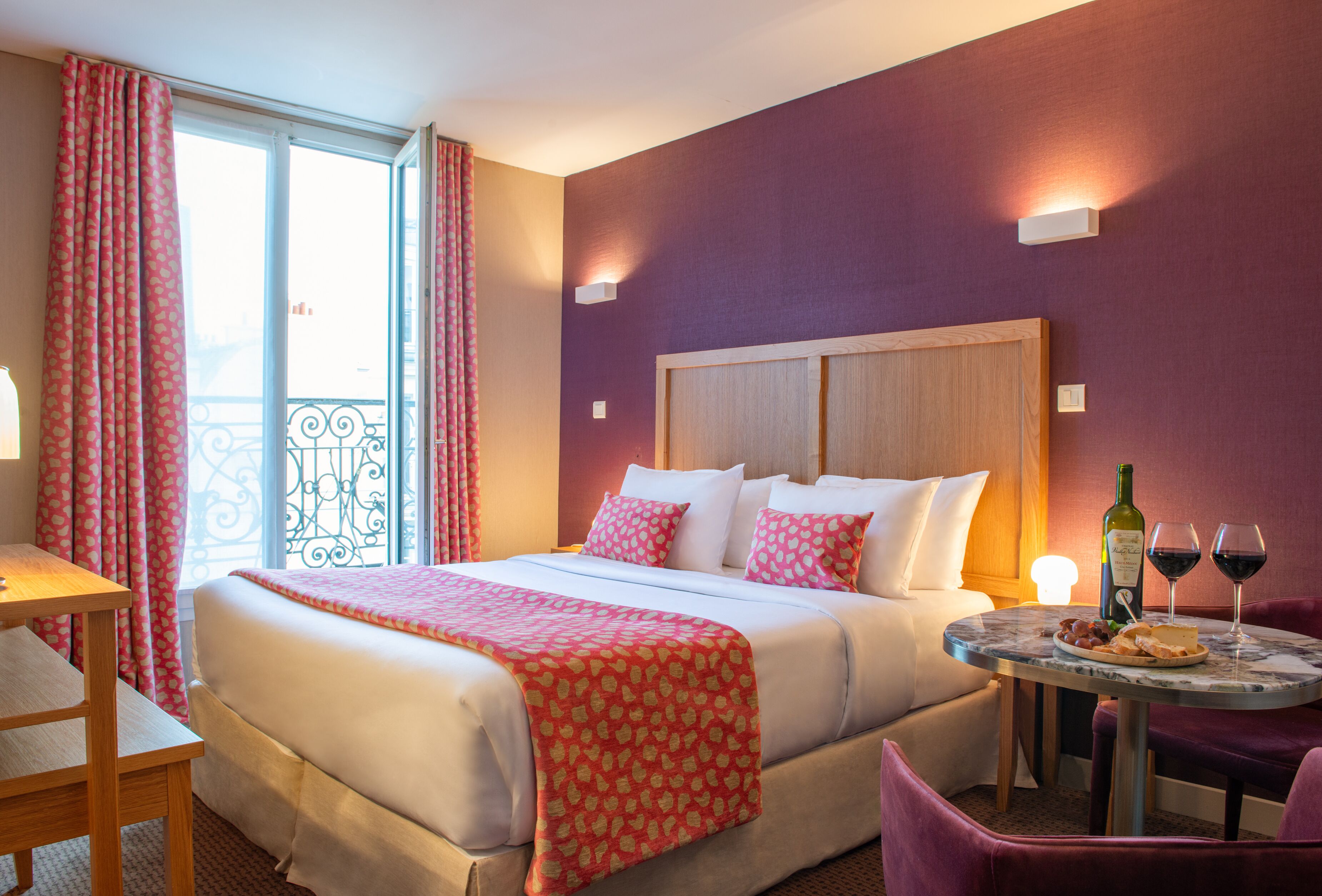 Habitación de hotel con una cama grande, una mesita con copas de vino y un balcón con cortinas.