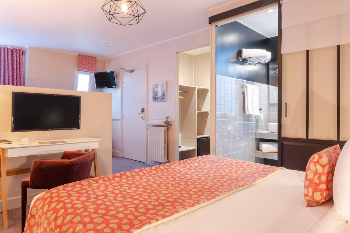 Habitación de hotel con cama, escritorio con televisor, colcha de cama roja estampada, una silla roja, baño con espejo y zona de ducha.