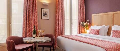 A hotel room with a bed, a table with two chairs, a bottle of champagne, and a framed picture on the wall.