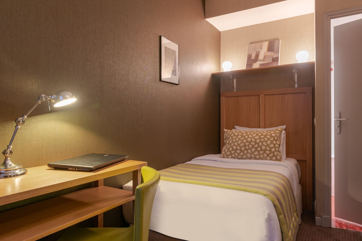 Habitación de hotel con una cama, un escritorio con una computadora portátil, una silla, una lámpara y una puerta.