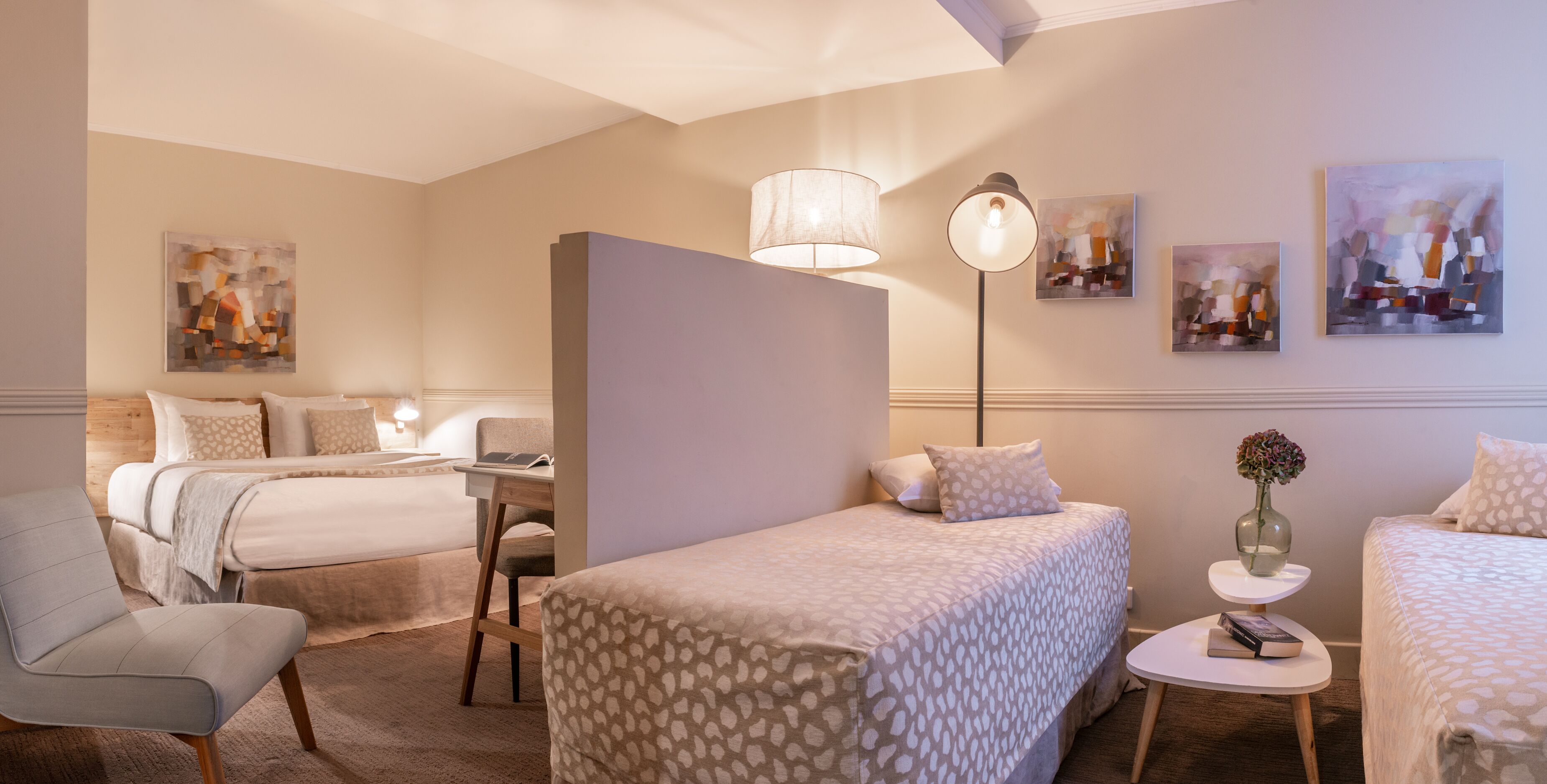 A hotel room with two beds, a desk, a chair, a lamp, and artwork on the walls.