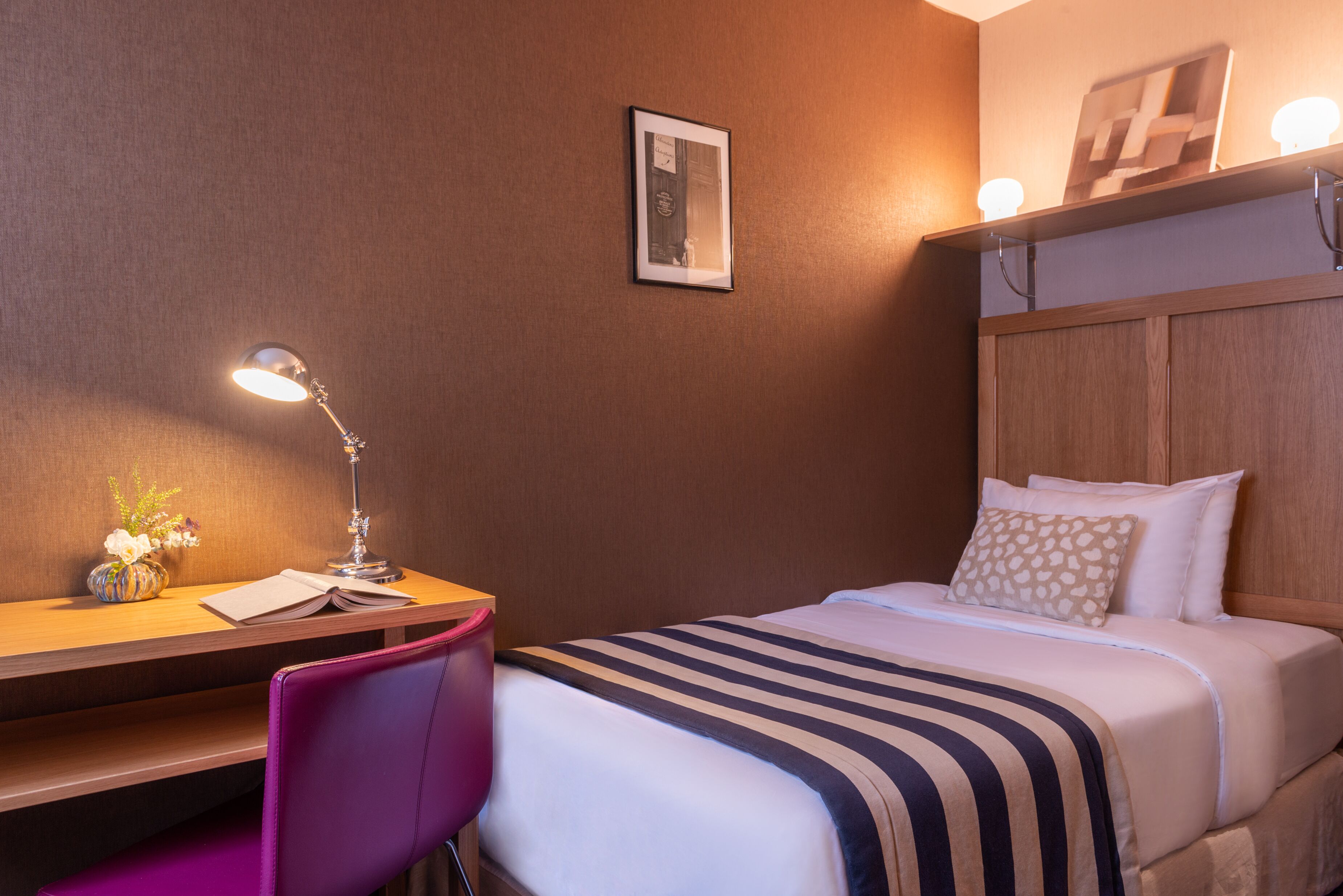 A hotel room with a bed, a desk with a lamp, a chair, and a small table with a vase of flowers.
